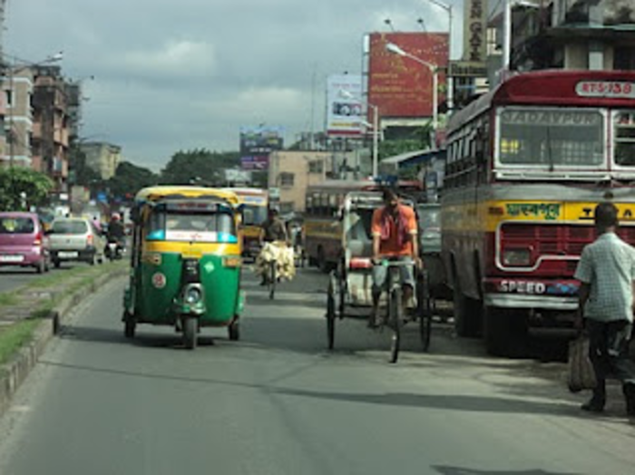 An auto rickshaw driving down the busy streets of Kolkata while a man carrying live chicken on his bicycle rides behind it