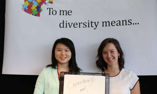Image of author standing beside another woman. They are standing in front of a black and white background which has the text "Diversity means..." written on a white with black ink.