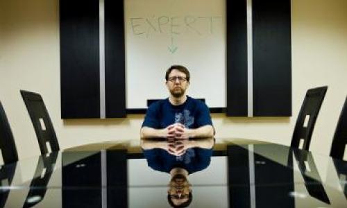 dave sitting at the head of a table with an arrow pointing at his head, with the words "expert"