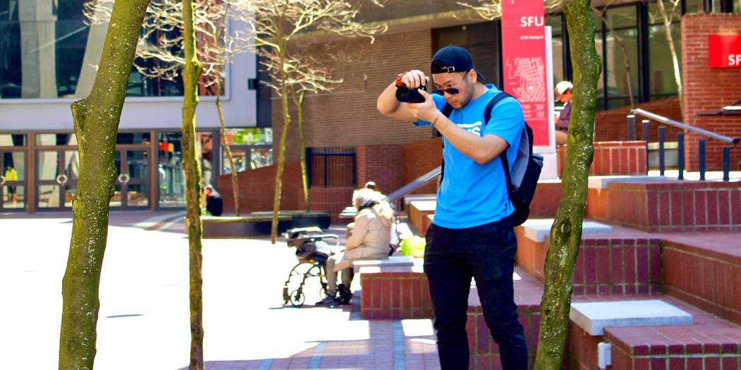Elvis standing in front of Simon Fraser University holding a camera