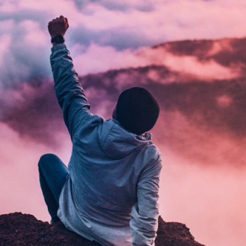person sitting on a cliff with their fist raised to their air in success