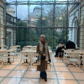 A young woman stands in a tiled cafe, with a window wall as her background. Beyond is a green courtyards surrounded by Romanesque buildings.