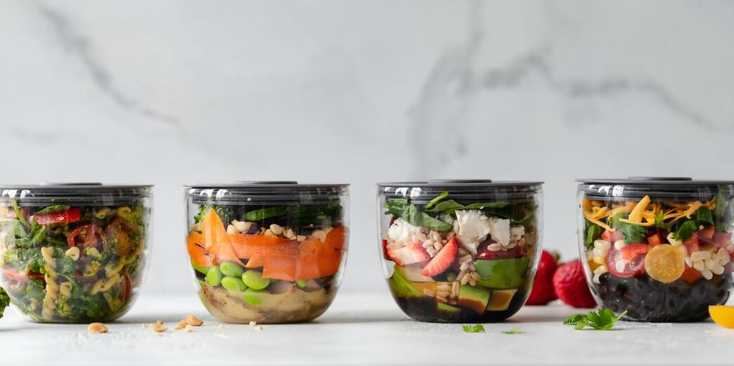 Row of four different food bowls containing (from left to right), pasta, salad, and two fruit salads