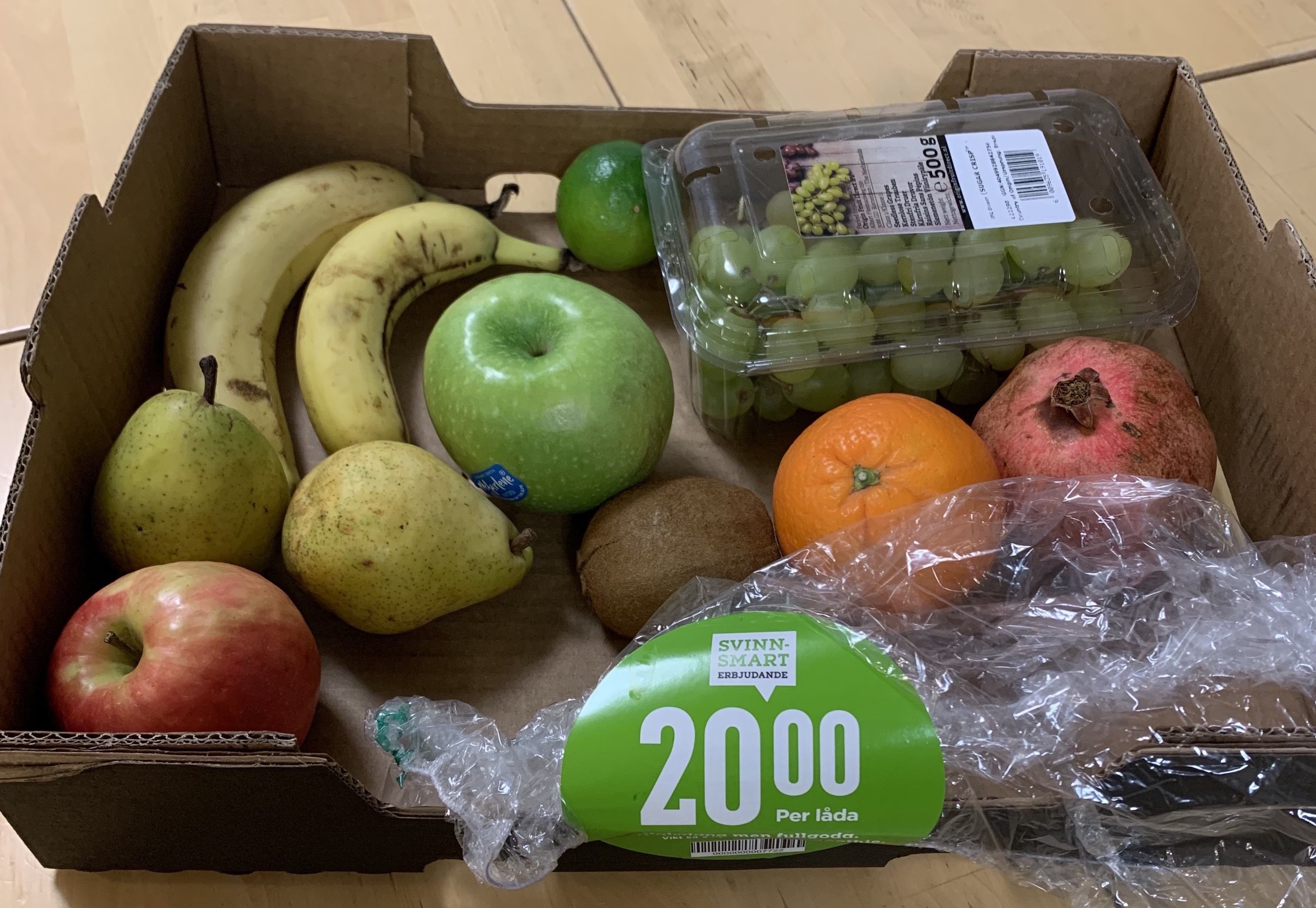 A cardboard box of discounted fruit, including bananas, apples, grapes, pomegranate, pear, and kiwi