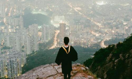 Guy wearing his graduation gown, standing at the edge of a cliff overlooking the city