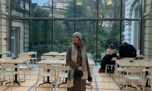 A young woman stands in a tiled cafe, with a window wall as her background. Beyond is a green courtyards surrounded by Romanesque buildings.