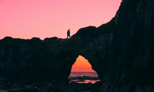 Image of a person walking on a rock arch over water during a pink sunset