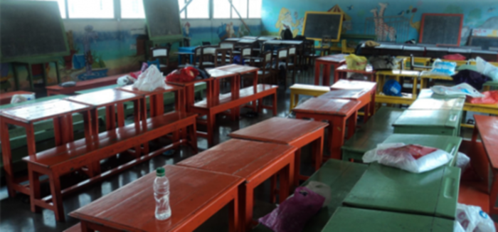 Table and chairs lined up in a classroom with classroom paraphernalia on the tables 
