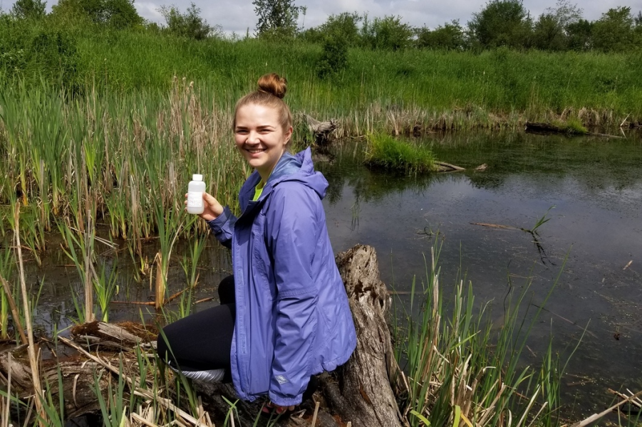 the author conducting a water sampling in Katzie Slough, holding a sample bottle and smiling at the camera