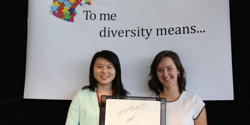 Image of author standing beside another woman. They are standing in front of a black and white background which has the text "Diversity means..." written on a white with black ink.