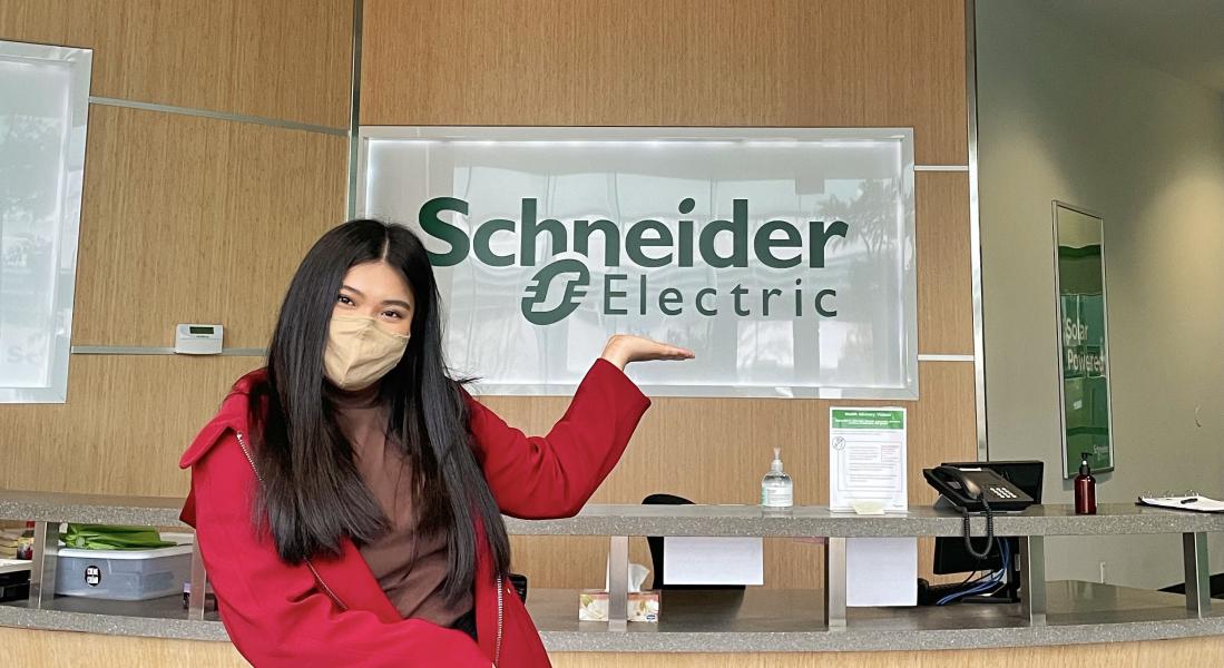 Girl standing next to sign that says "Schneider Electric", pointing at sign.