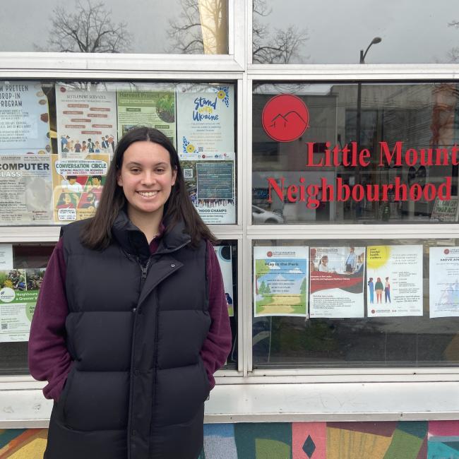 Jordan standing in front of Little Mountain Neighbourhood House's window with several posters of available programs that the house offers
