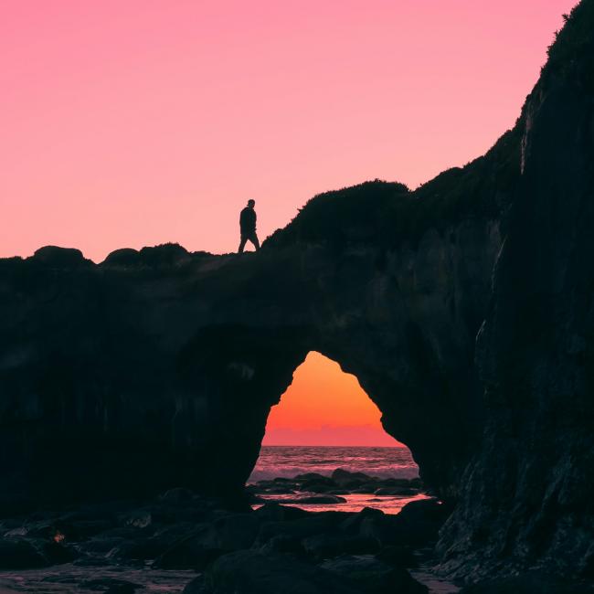 Image of a person walking on a rock arch over water during a pink sunset