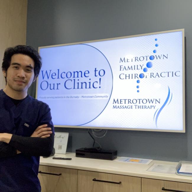 Ardie Dizon standing in front of a screen with text that says "Welcome to Our Clinic!" and logos for Metrotown Family Chiropractic and Massage