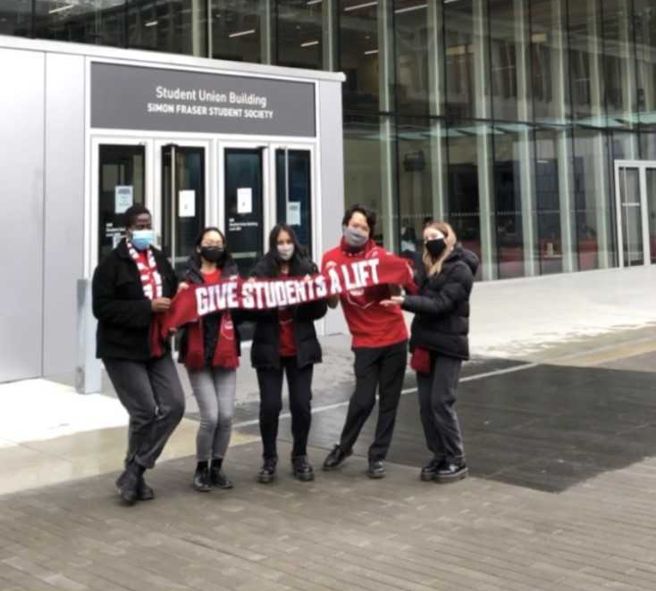 Five students holding a banner that says "Give students a lift" in front of the library at the SFU Burnaby campus