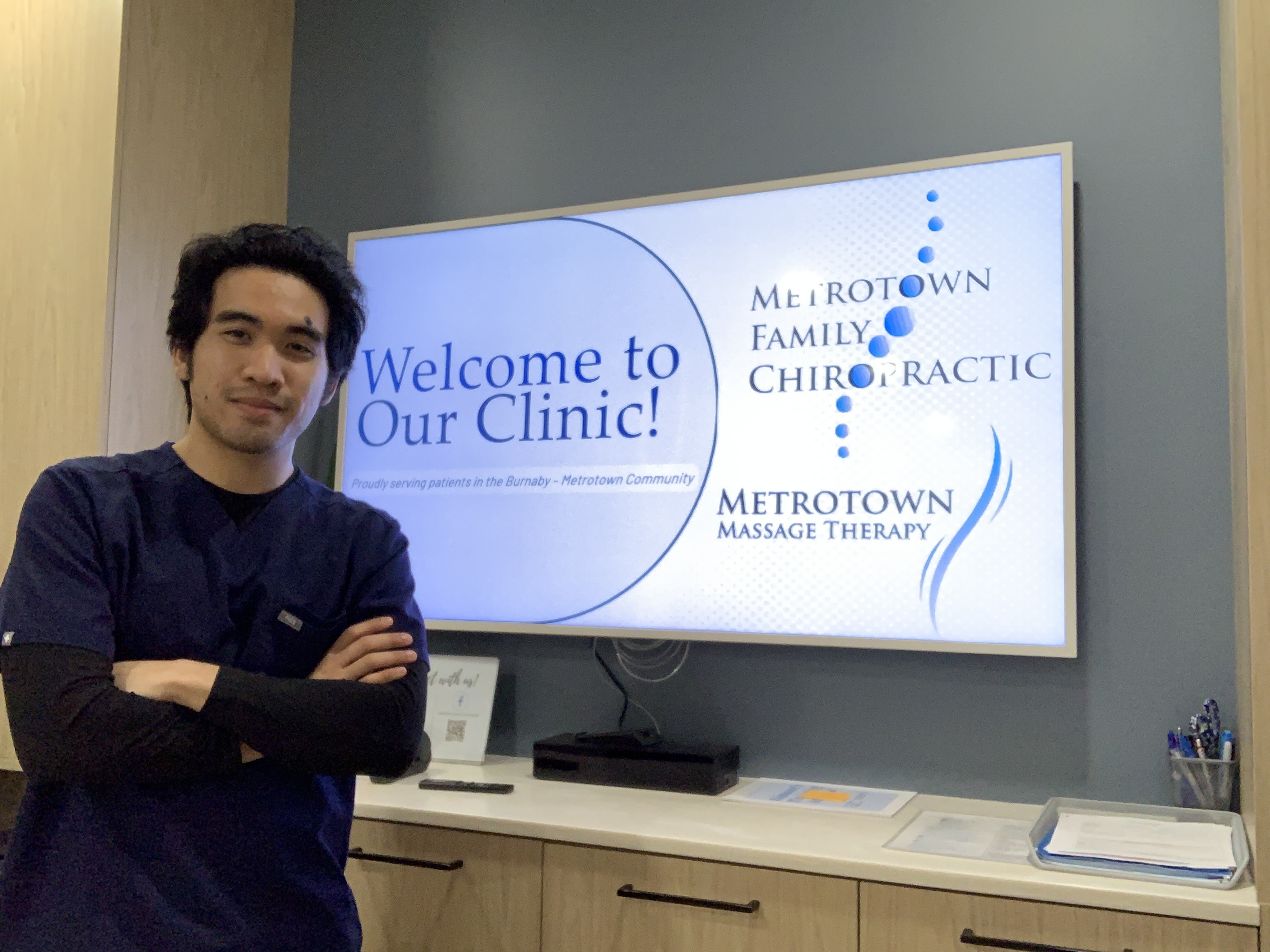 Ardie Dizon standing in front of a screen with text that says "Welcome to Our Clinic!" and logos for Metrotown Family Chiropractic and Massage