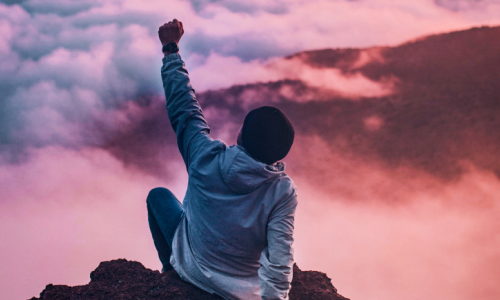 person sitting on a cliff with their fist raised to their air in success