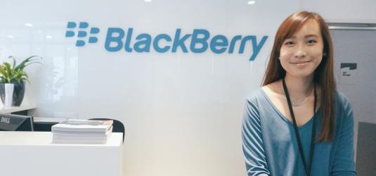 Image of Author. She is smiling at the camera and is wearing blue blouse. The wall behind her has the logo of Blackberry on it.
