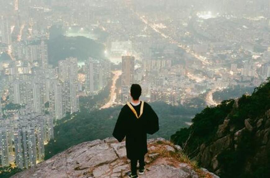 Guy wearing his graduation gown, standing at the edge of a cliff overlooking the city