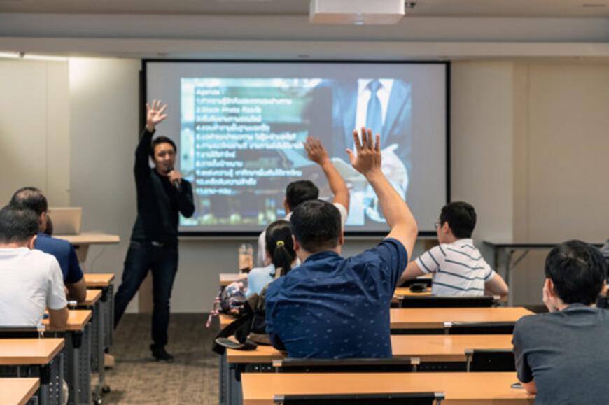 Man standing in front of room giving a presentation while his audience raise their hands up to answer a question
