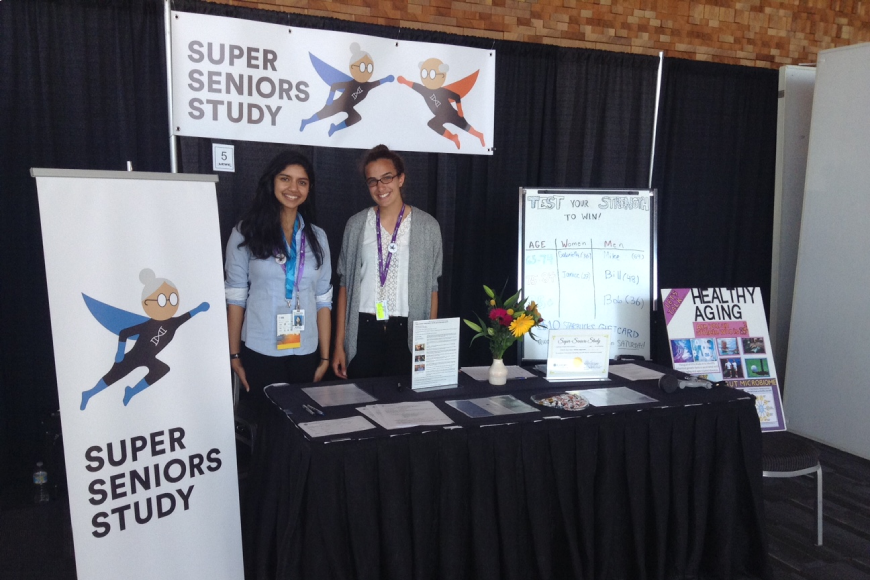 Both authors standing in a booth showcasing their research with posters of super seniors in comic form, around them.