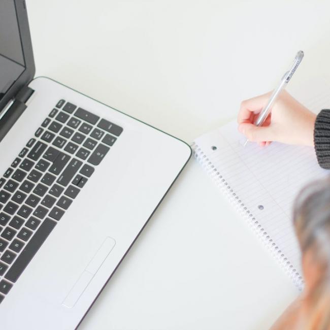 A person writing on paper with a laptop on the desk