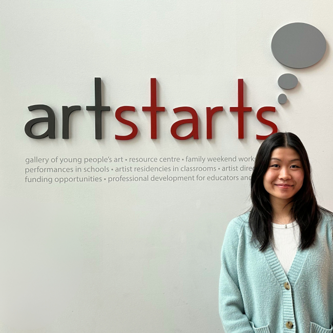 Erin Lum standing in front of a white wall with the logo for ArtStarts in Schools.