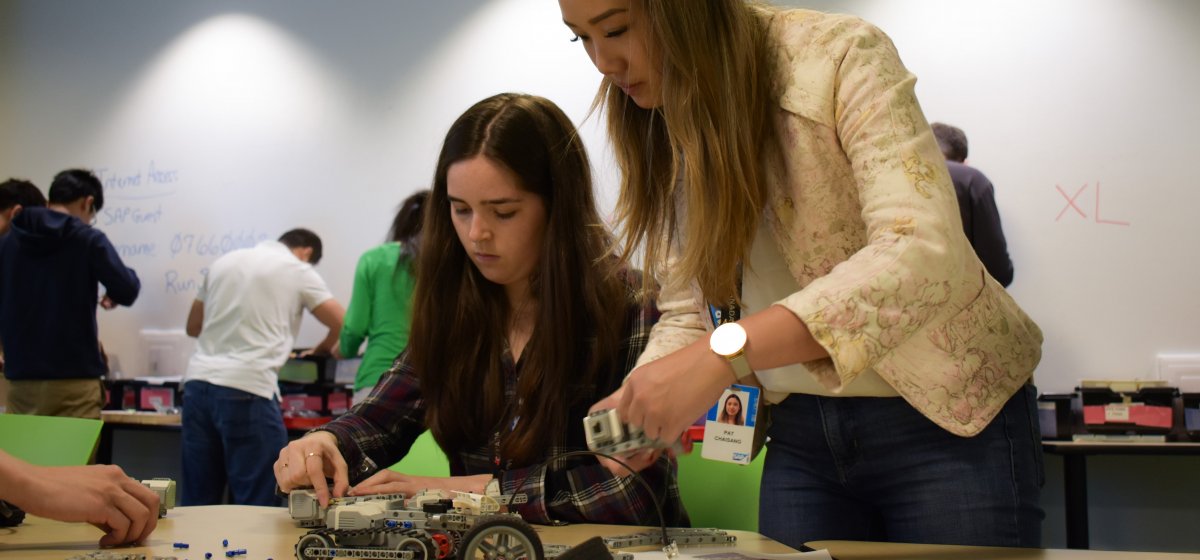 Two females student are working on a prototype in a lab with a few other students standing behind.