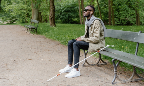 A young man sitting on a park bench, listening to music with his seeing cane resting beside him