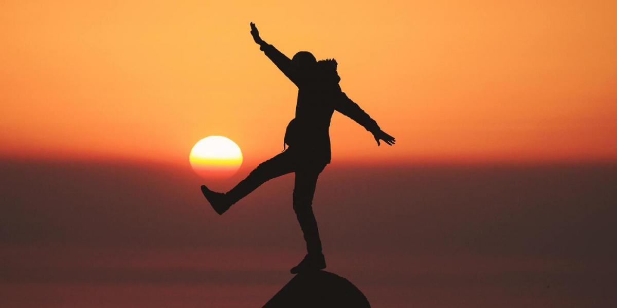 Person standing on one foot on rock, in front of sunset 