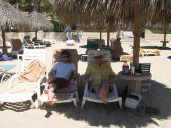 Dave and his father sit on lounge chairs at the beach. A straw umbrella shades them from the sun.