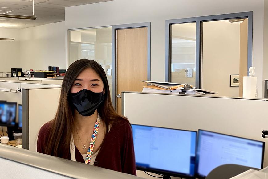 A young women with a mask is standing in her work cubicle with a computer behind her. 
