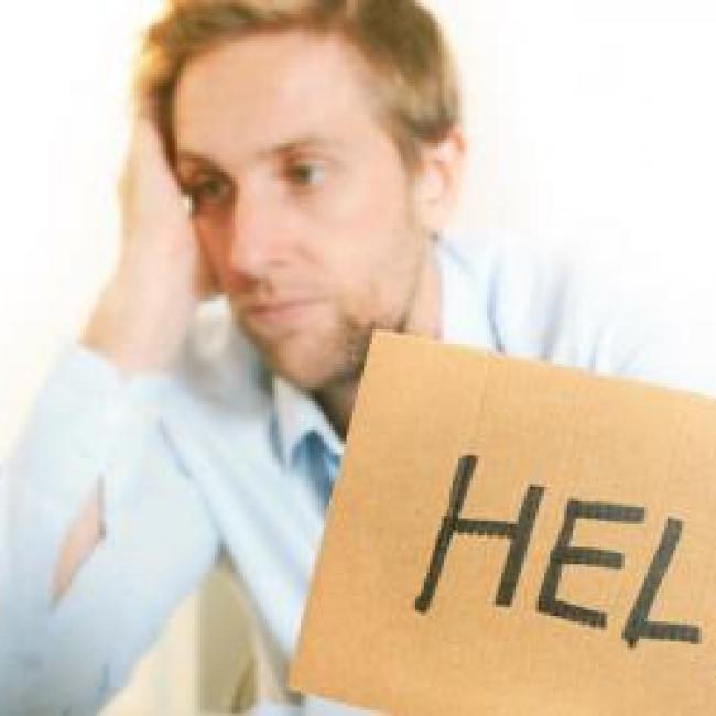 A man staring at a laptop screen. He looks frustrated and has a cardboard piece in his hand with the word help written on it.