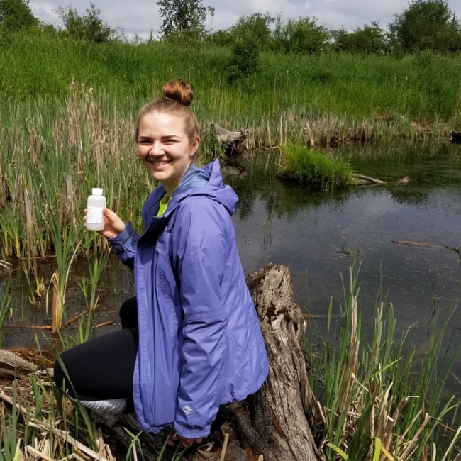 the author conducting a water sampling in Katzie Slough, holding a sample bottle and smiling at the camera
