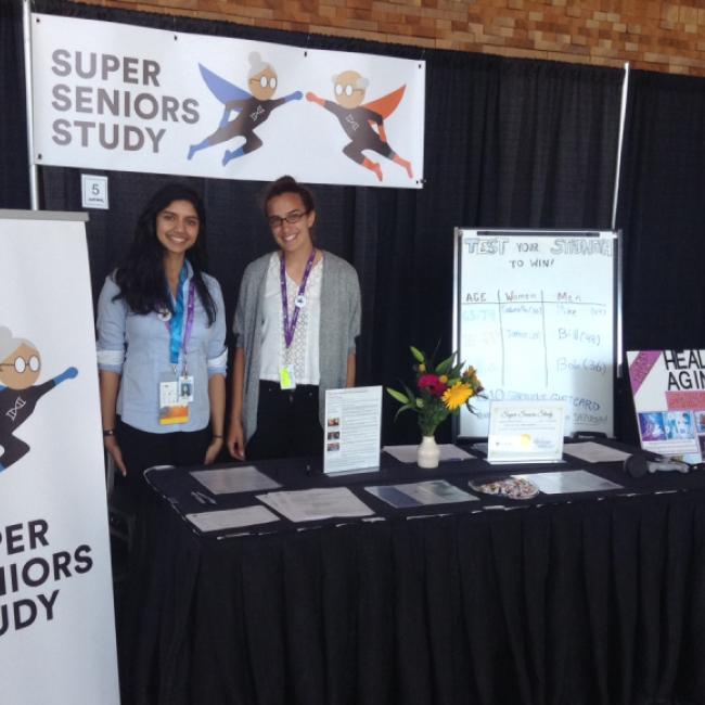 Both authors standing in a booth showcasing their research with posters of super seniors in comic form, around them.