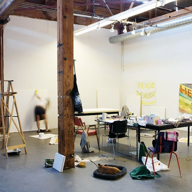 Image of an art workshop. It is a big room with wooden pillars and roof beams. There is a wooden ladder in the room. Some tables and chairs and some paintings on the walls.