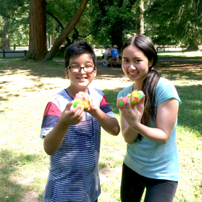Author and her young male student smiling at the camera and holding up tennis balls in both hands.The picture is set in a field with trees in the background.