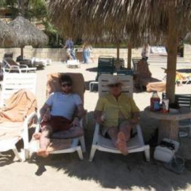 Dave and his father sit on lounge chairs at the beach. A straw umbrella shades them from the sun.