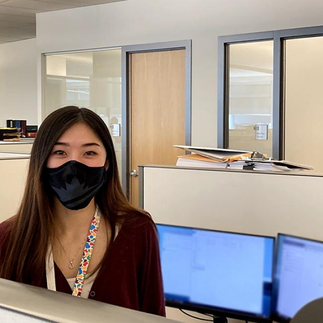 A young women with a mask is standing in her work cubicle with a computer behind her. 