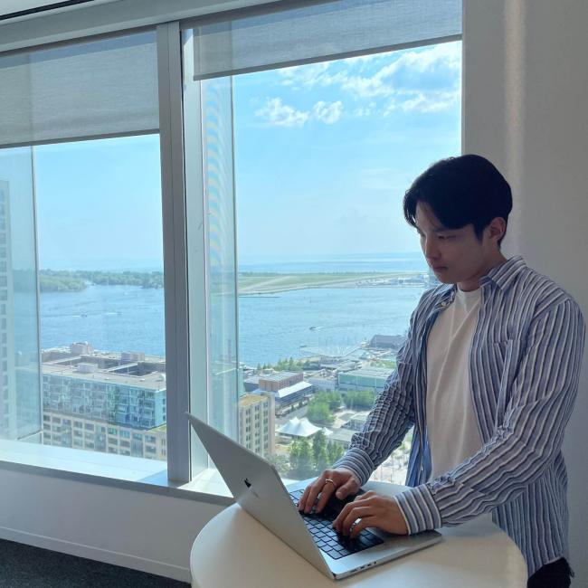 Josh Kim standing by a window with a view while working on a laptop