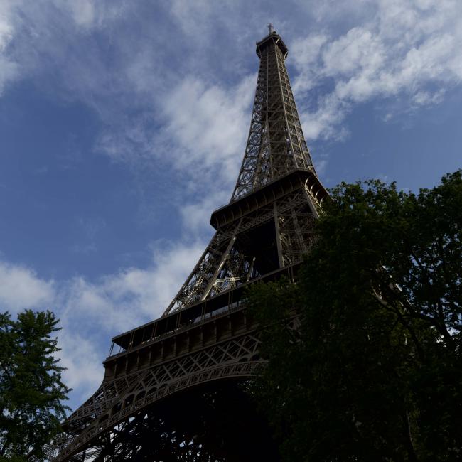 Photo of the Eiffel Tower taken from below. The sky behind is blue with white clouds.